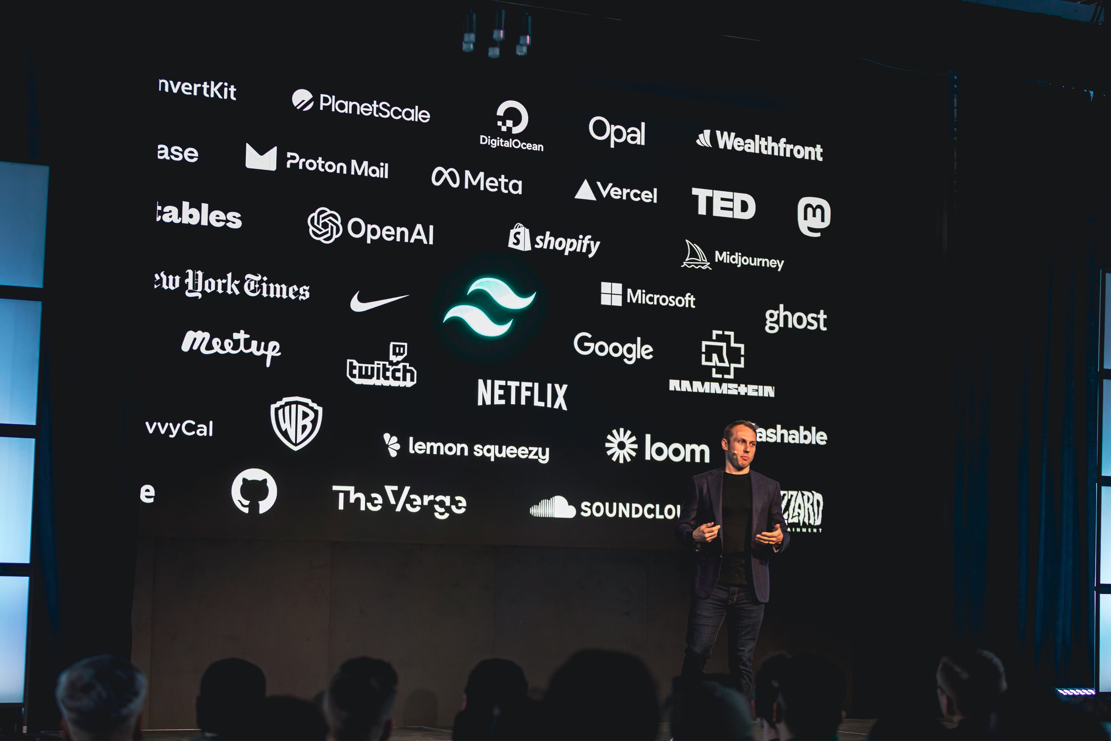 Adam Wathan de pie en el escenario frente a una diapositiva que destaca algunas de las empresas que usan Tailwind CSS